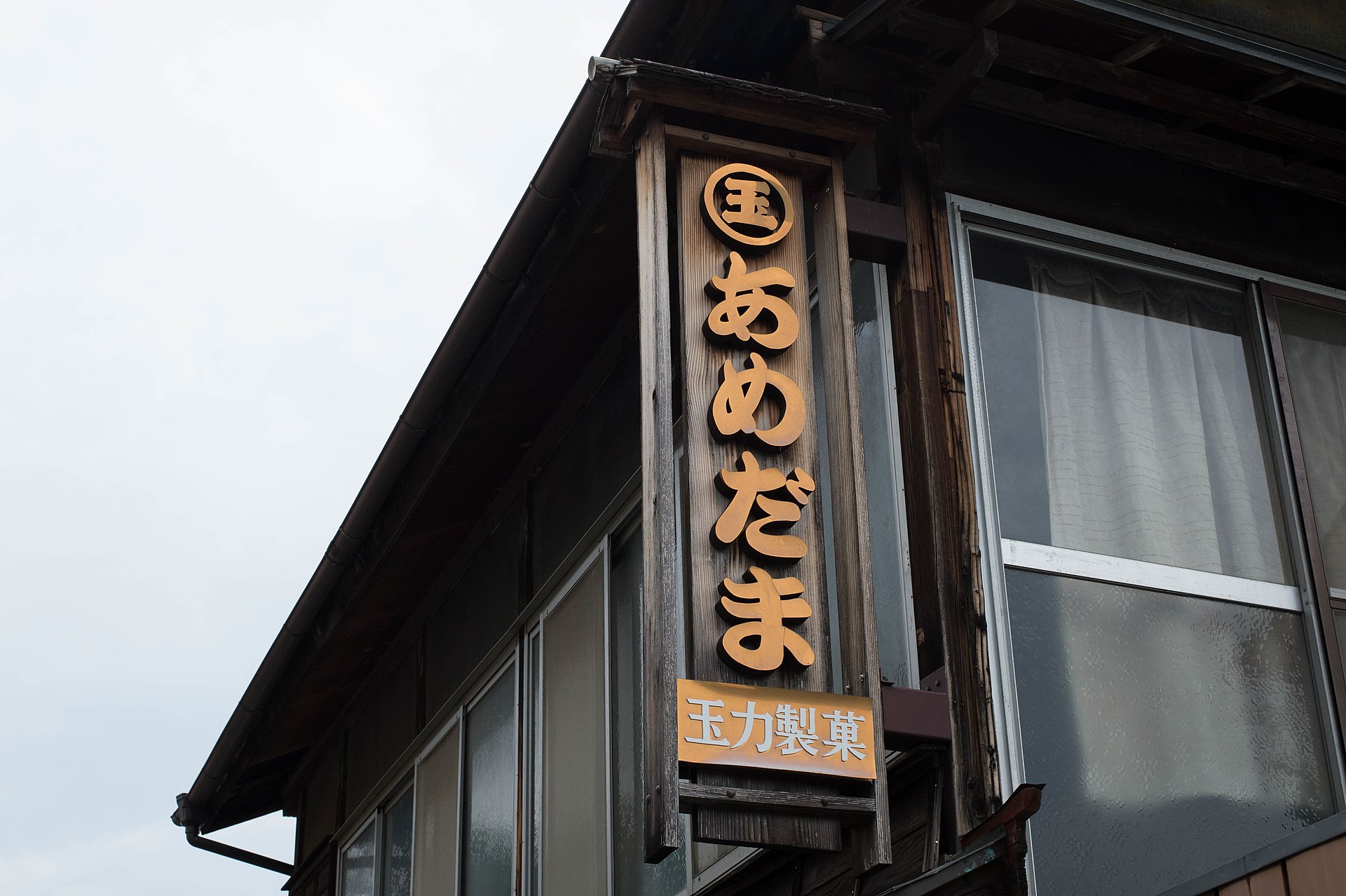 仰拍旧木造店铺的屋檐一角,突出的竖长木招牌上有金色假名「あめだま」,下方小牌写着「玉力製菓」。