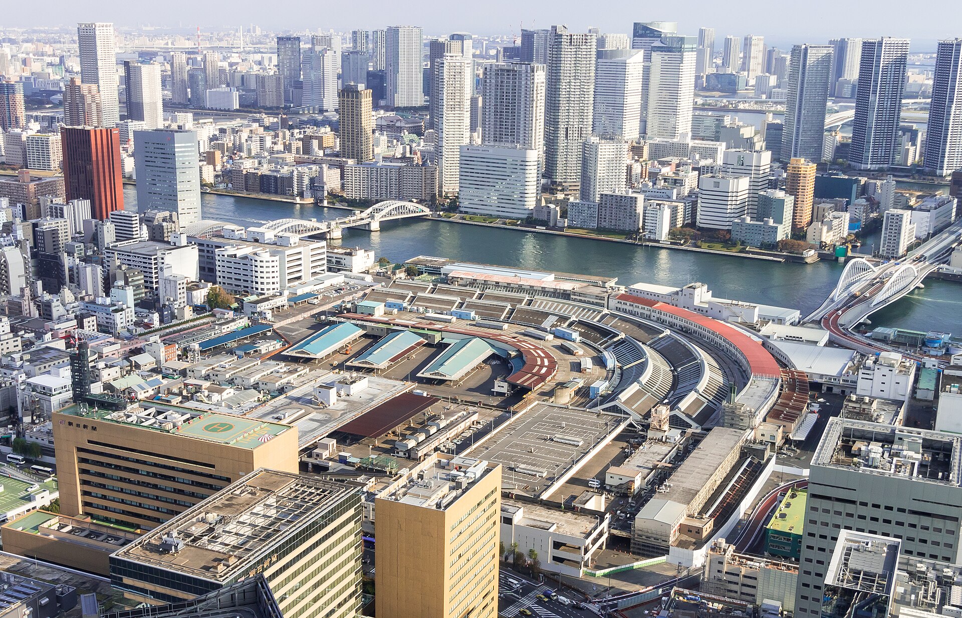 从高处俯瞰筑地市场扇形排列的屋顶群,远处是隅田川、桥梁与东京的高楼天际线。