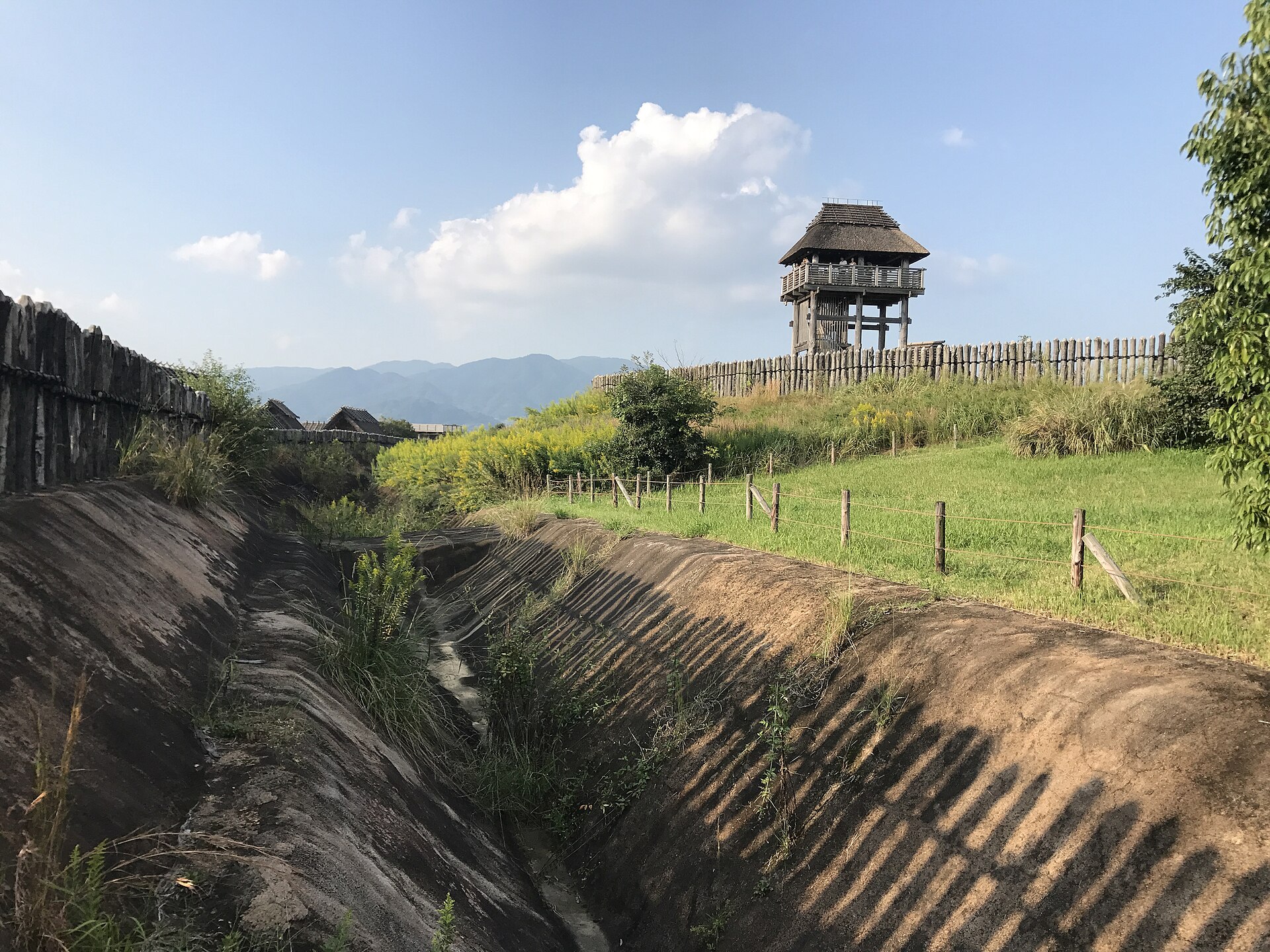 V字形壕沟与木栅栏，土丘上矗立瞭望楼，远处群山连绵。