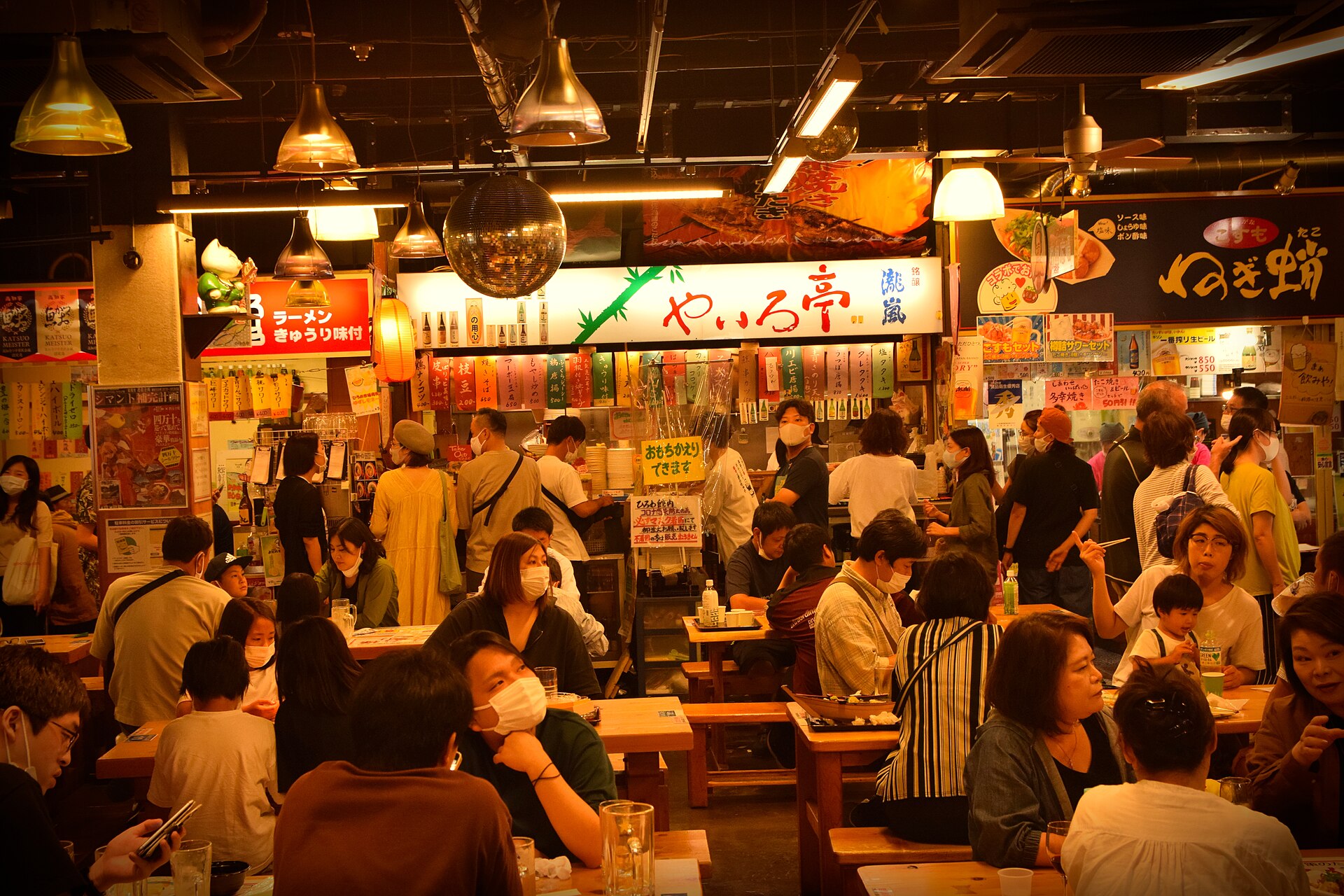 高知弘人市场室内，人们围坐在长木桌旁用餐，身后是亮着灯的日式美食摊位。