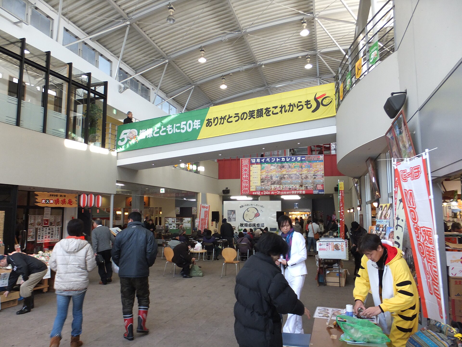 秋田市民市场室内通道，钢桁架天花板下悬挂着五十周年横幅，购物者在摊位之间走动。