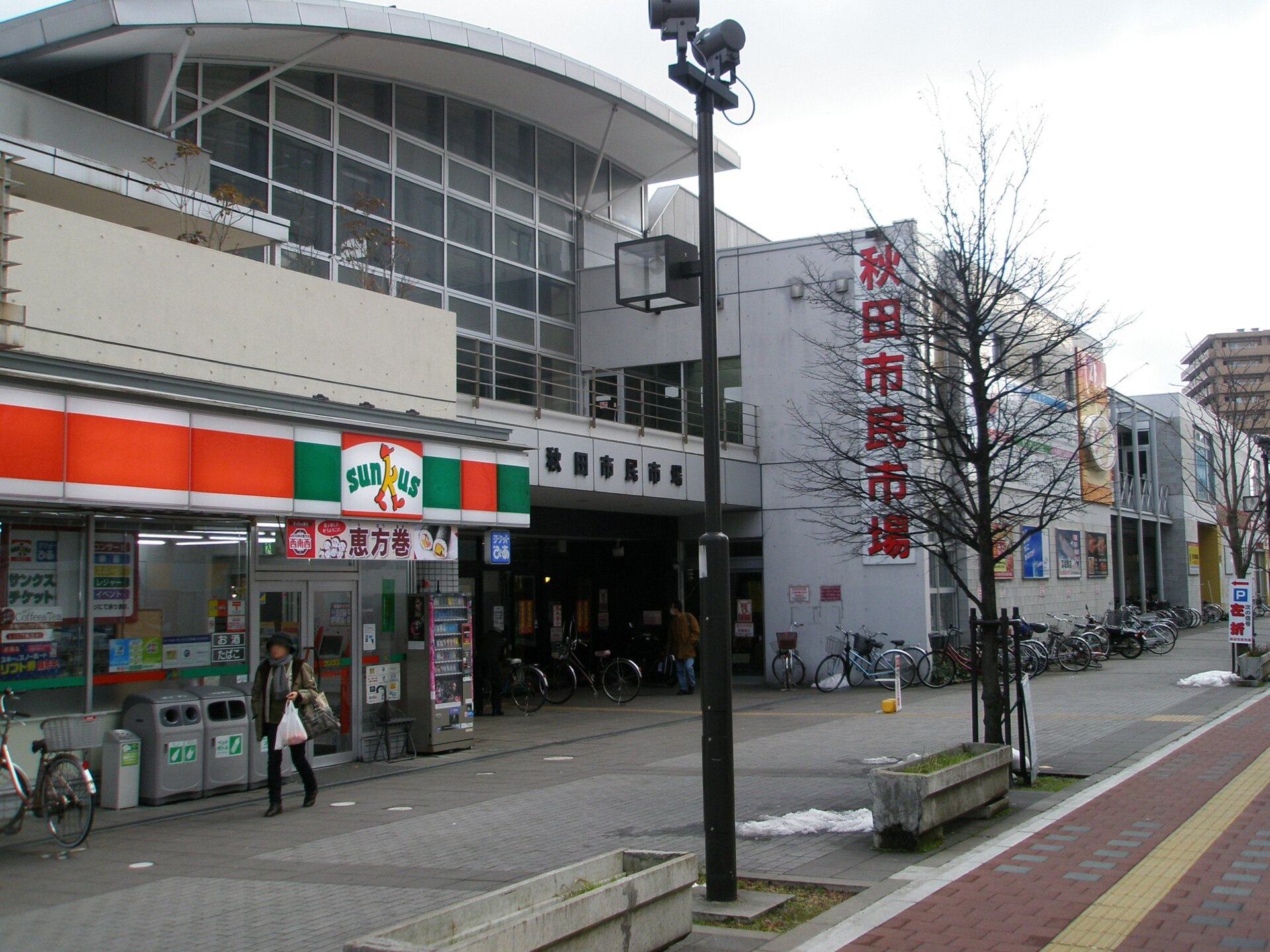 秋田市民市场的临街外观，白墙上是红色竖排招牌，楼前停着成排自行车，左侧为一家便利店。