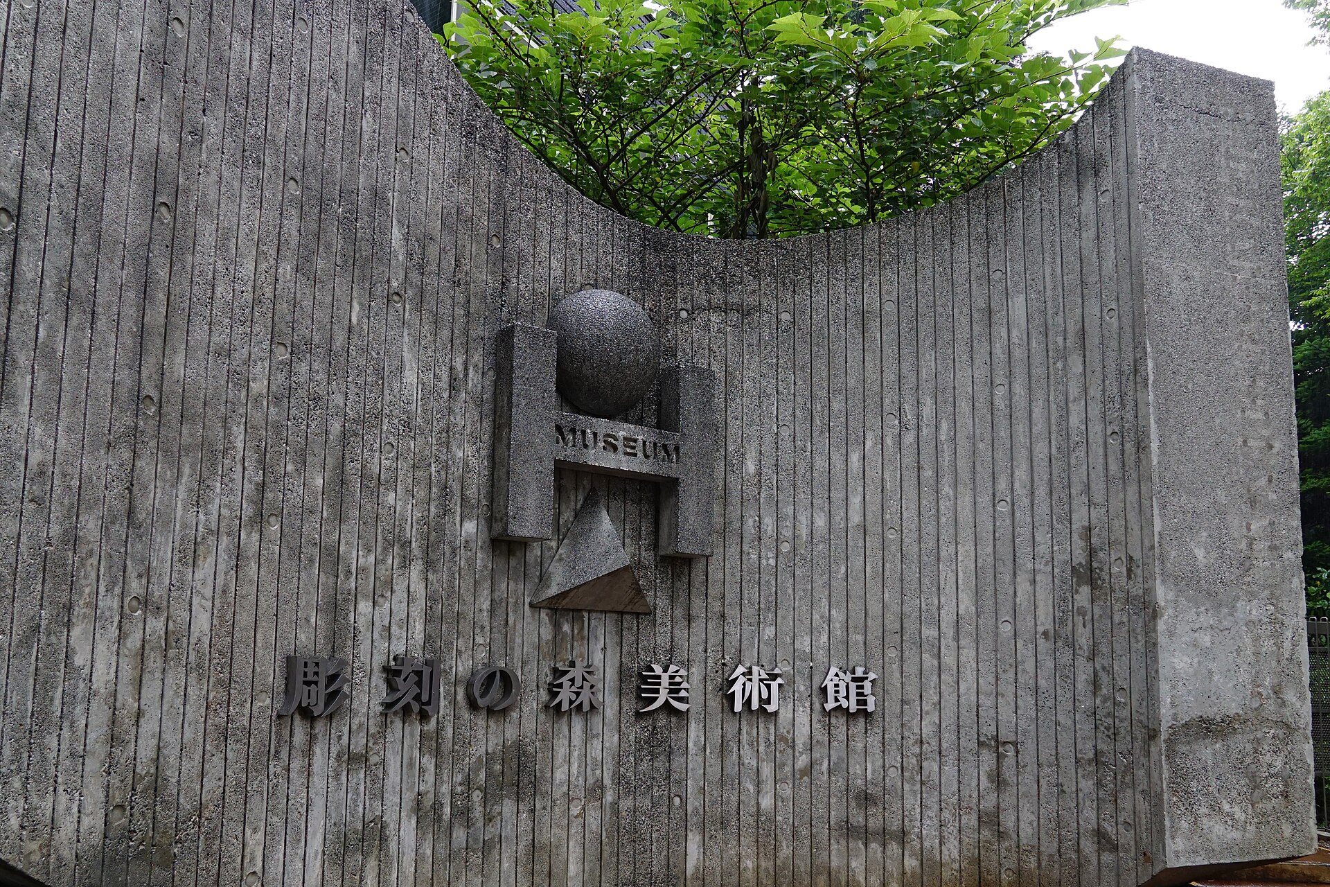 仰拍箱根雕刻之森美术馆入口的竖纹混凝土墙，墙面嵌着球体浮雕标志与馆名金属字，弧形缺口上方探出绿叶。