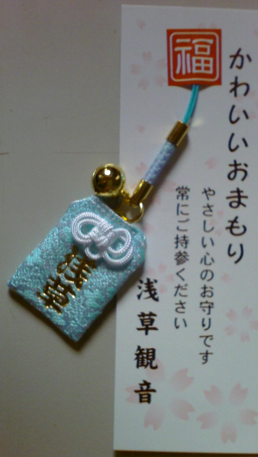 一枚浅蓝色的浅草御守，带金色小铃，与印有樱花的说明卡一起放在浅色桌面上。