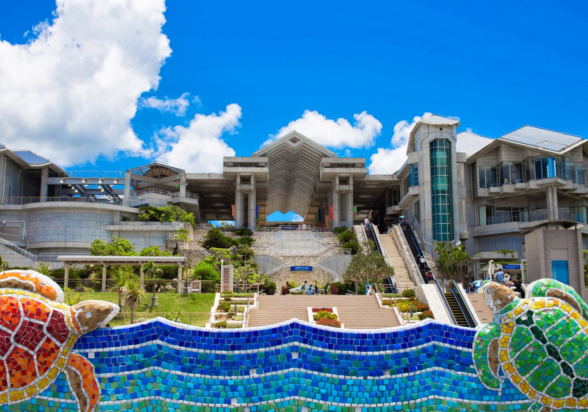 蓝天白云下的冲绳美丽海水族馆建筑外观，前景是波浪形马赛克墙与海龟雕塑。