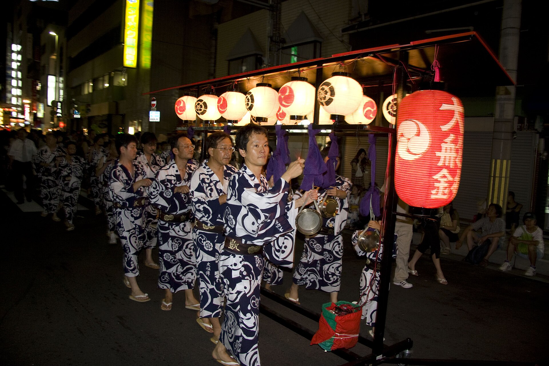 夜晚街头，一列身着蓝白浴衣、手持小钹的乐手行进，旁边灯架上挂着白纸灯笼和写有“大船鉾”的红灯笼。