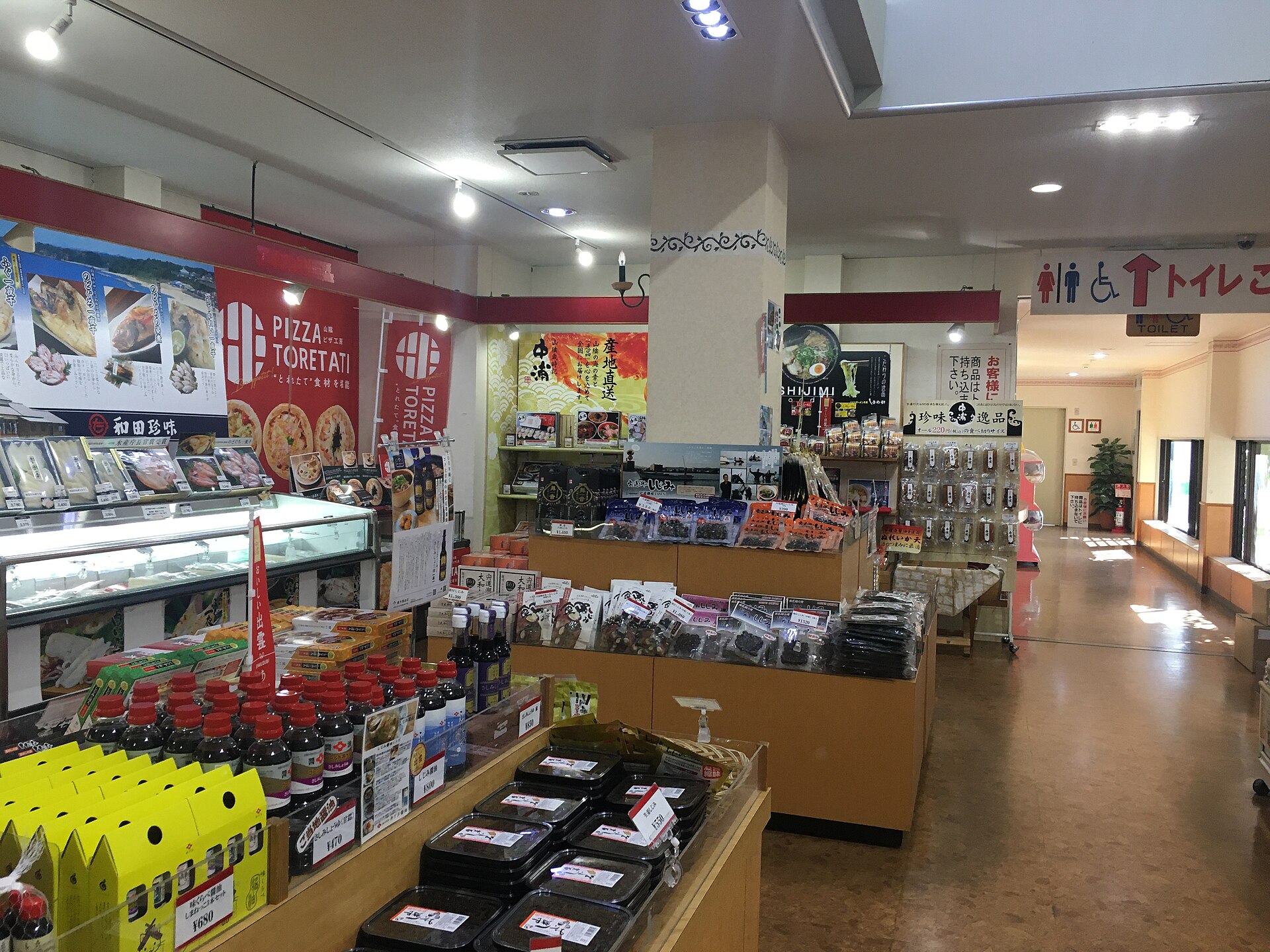 店内土特产卖场一角，木质货架上摆放着包装海产、成排的红盖酱油和黄色礼盒，左侧是冷藏陈列柜。