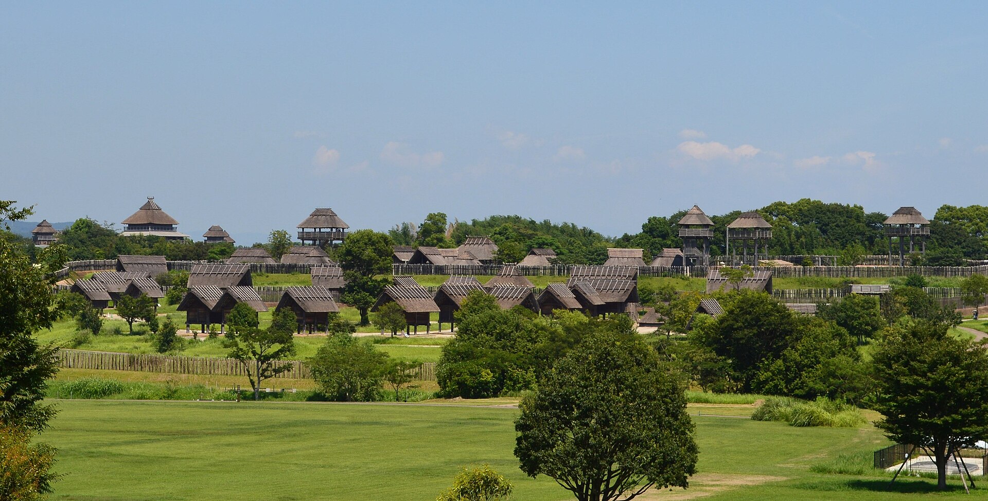 复原弥生聚落全景，草地对面排列着干栏仓库、竖穴住居与瞭望楼。